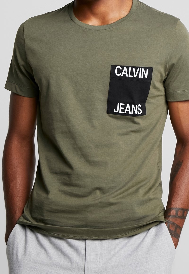 Mies, joka ei yllään oliivinvihreä t-paita, jossa on musta rintatasku, johon on merkitty "Calvin Jeans". Hänen kätensä ovat vaaleanharmaiden housujen taskuissa, ja hänellä on näkyvä käsivarttattoos.