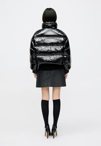 Μαύρο κοντό πούφο μπουφάν με γυαλιστερή φινίρισμα, οριζόντιο καπιτονέ σχέδιο, ψηλό γιακά και ελαστικές μανσέτες, συνδυασμένο με τζιν φούστα.