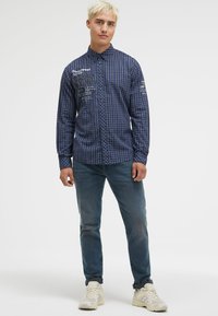 Dunkelblau kariertes Hemd mit Button-Down-Kragen, versehen mit weißen Textgrafiken, kombiniert mit hellblauen Jeans und beigen Sneakern.