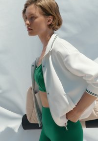 Jeune femme en tenue de sport verte et veste blanche zippée debout de profil devant un fond clair et texturé.