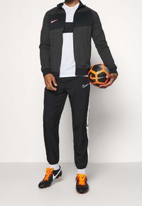 Mann in schwarz-grauem Nike Trainingsanzug, der einen schwarz-orangenen Nike Fußball hält, trägt schwarze Fußballschuhe mit leuchtend orangenen Schnürsenkeln und weiße Socken.