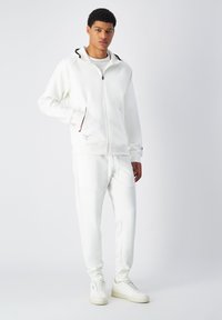Felpa bianca con zip e accenti neri, abbinata a pantaloni bianchi coordinati. Entrambi i capi presentano una trama liscia e un design sportivo.