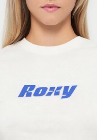 White cotton t-shirt featuring a prominent blue graphic logo "Roxy" across the chest in a bold, stylized font.