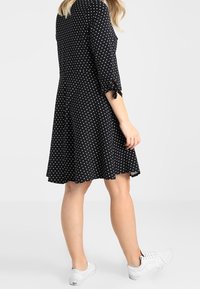 Černé šaty s puntíky, tříčtvrtečními rukávy a mašlí na manžetách. Plynulý střih do áčka. Nošeno s bílými teniskami.
