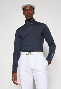 Granatowa koszula polo z długim rękawem i kołnierzykiem na zamek błyskawiczny, w połączeniu z białymi spodniami i białym paskiem z metalową klamrą, noszona przez mężczyznę.