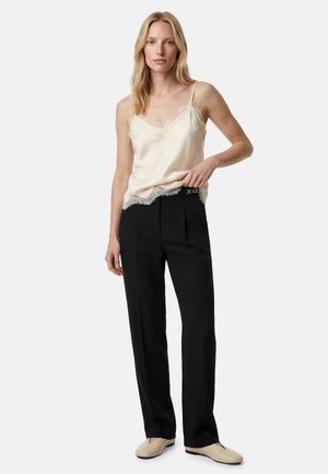 Frau trägt ein beiges Spitzen-Camisole, schwarze hoch taillierte Hose und beige flache Schuhe, steht mit einer Hand in der Hüfte.