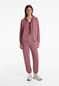 Übergrößiger, mit Reißverschluss versehener Kapuzenpullover und passende Jogginghose in einem hellen Mauve-Ton. Weicher Stoff, elastische Bündchen und kontrastierende Stoffdetails.