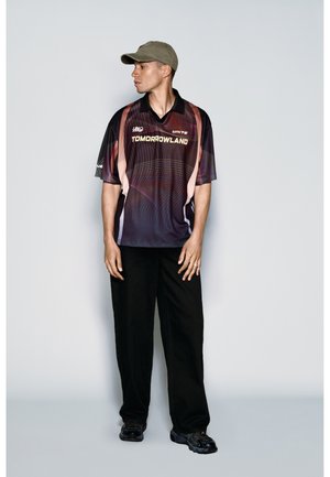 Junger Mann trägt ein schwarz-pinkes Tomorrowland-Shirt, schwarze Hose, schwarze Sneakers und eine olivgrüne Baseballkappe, steht vor einer grauen Wand.