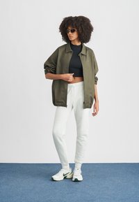 Ελαφρύ τζάκετ σε ελιά πράσινο, μαύρο κοντό τοπ, γκρι φόρμα, και λευκά αθλητικά παπούτσια με πράσινες λεπτομέρειες· μοντέλο με γυαλιά ηλίου.