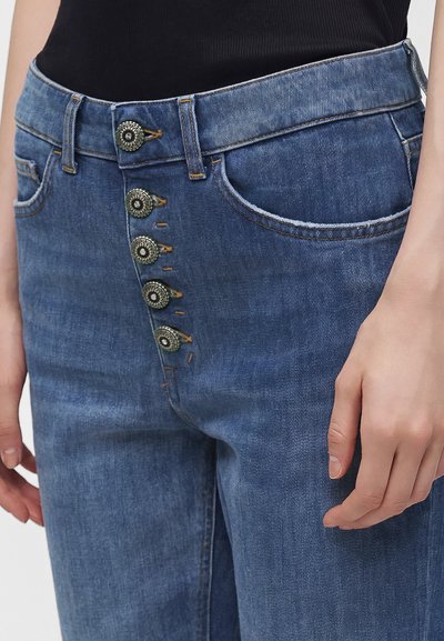 Gros plan d'un jean en denim bleu avec cinq boutons argentés décoratifs le long de la braguette et la main d'une personne reposant à côté.