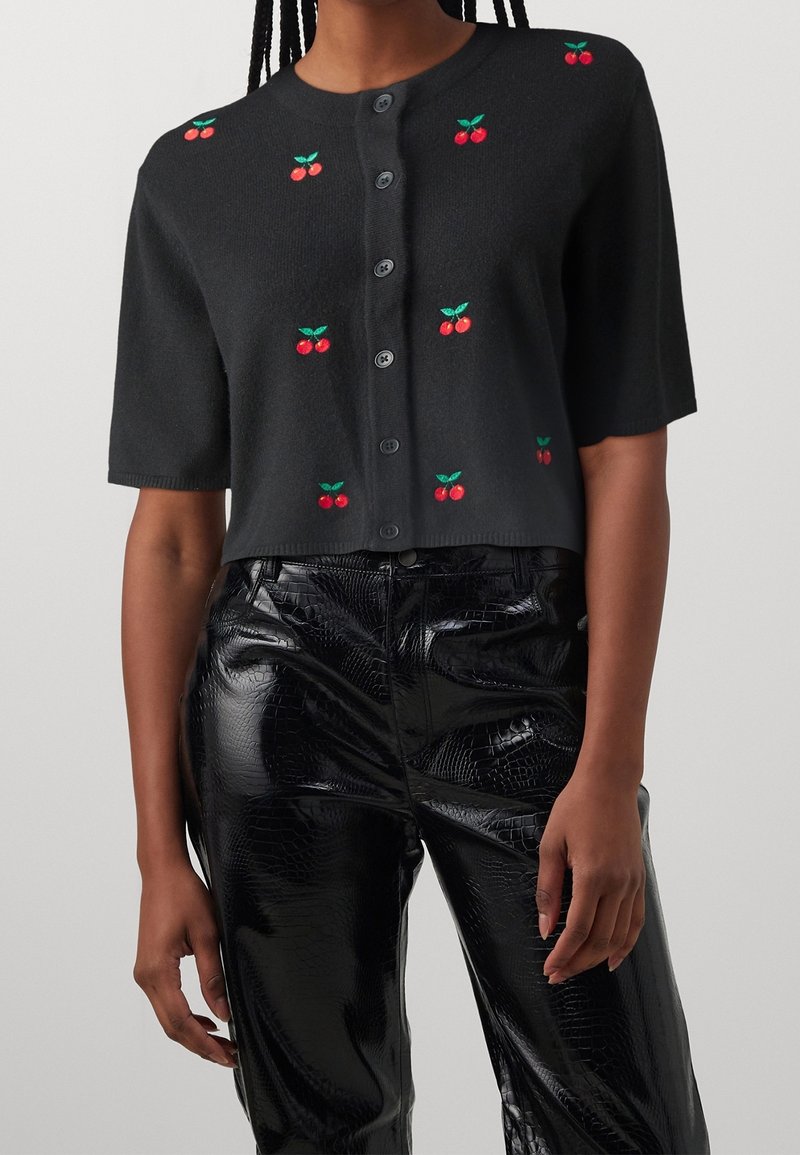 Cardigan noir à manches courtes boutonné avec une petite broderie de cerises rouges, porté avec un pantalon noir brillant texturé.