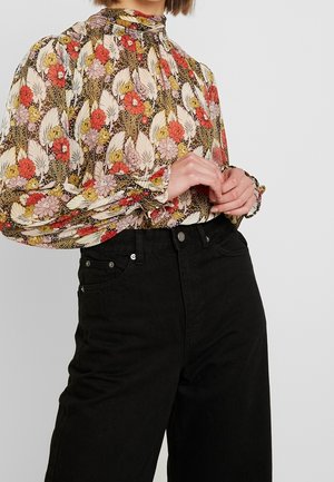 Persona con una blusa de manga larga con flores rojas, amarillas y rosas, metida dentro de unos vaqueros negros de talle alto, con las manos entrelazadas.