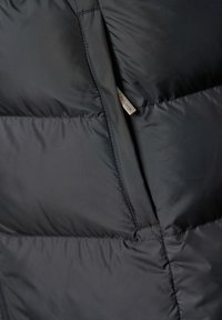 Svart puffer-jakke med quiltet tekstur og glidelåslommer. Har en liten etikett med merkevarenavn nær sømmen. Mykt, lett materiale.
