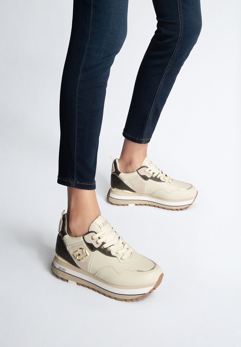 Femme portant des baskets compensées crème et marron avec des lacets beiges, associées à un jean skinny bleu foncé sur un fond uni.