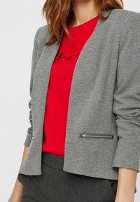 Grå blazer med en struktureret finish, der har reverskrave, åben front og lynlåslommer i siden. Båret sammen med en rød T-shirt.