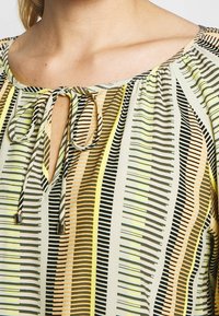 Blouse avec un décolleté en forme de trou de serrure et un nœud, fabriquée dans un tissu léger aux rayures vertes, jaunes, noires et pêche. Motif de lignes verticales.