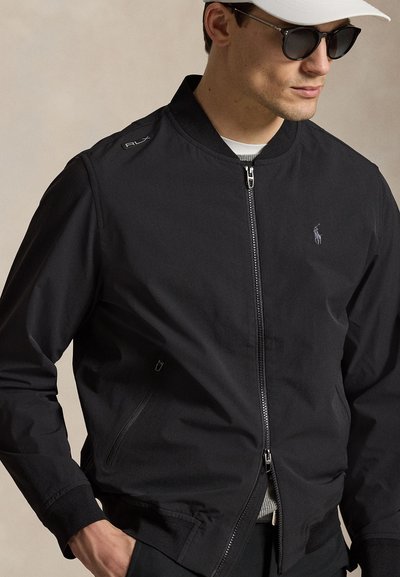 Muž nosící černou bundu na zip s logem Ralph Lauren, bílou čepici a černé sluneční brýle, pózující na jednobarevném pozadí.