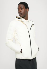 Polstret jakke i off-white med sort lynlås og detaljer. Har hætte og skrå quiltet tekstur, parret med en lysefarvet skjorte.