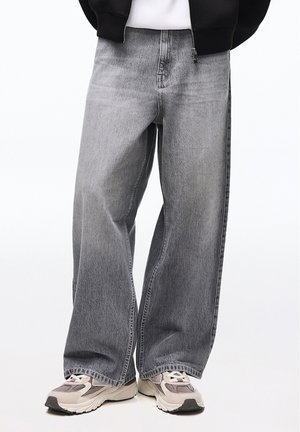 Person iført løstsiddende grå jeans og beige sneakers, stående med benene let spredt mod en ensfarvet hvid baggrund.