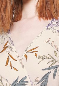 Tela ligera con estampado floral y escote en V. Presenta tonos de hojas verdes, marrones y moradas sobre un fondo beige.
