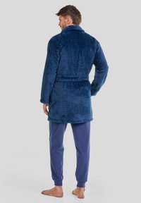 Bata de baño de terciopelo azul marino, con un patrón de cuadrícula texturizado y cuello chal, ajustada en la cintura con un cinturón a juego. Combinada con pantalones azul claro.