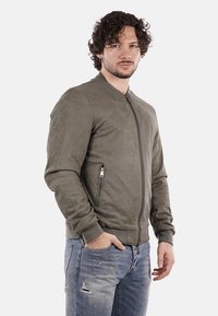Giacca bomber verde in suede con zip frontale, polsini a coste e due tasche laterali con zip. Indossata con jeans blu con effetto usurato.