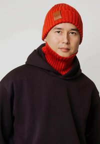 Czerwona dzianinowa czapka z prążkowaną fakturą i skórzaną naszywką z logo, zestawiona z pasującym czerwonym prążkowanym kominem na szyję. Noszona na ciemnym kapturze.