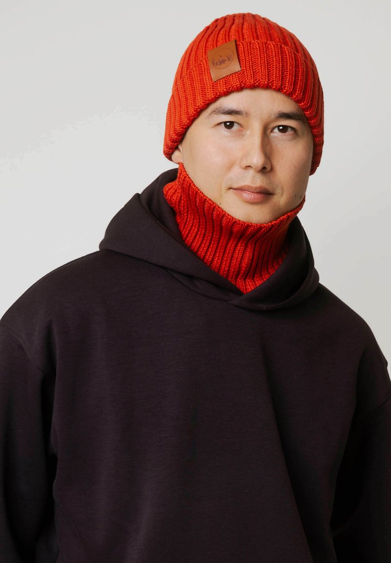 Czerwona dzianinowa czapka z prążkowaną fakturą i skórzaną naszywką z logo, zestawiona z pasującym czerwonym prążkowanym kominem na szyję. Noszona na ciemnym kapturze.