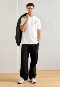 Witte katoenen t-shirt met een kleine grafische afbeelding op de borst, gecombineerd met losse zwarte cargobroeken en grijze sneakers met zwarte accenten.