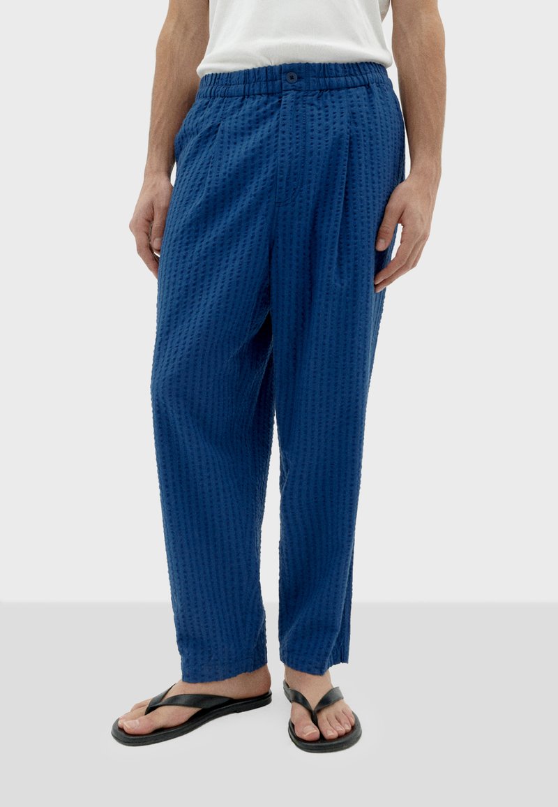 Blauwe textuurbroek met een elastische tailleband en knoopdetail; relaxte pasvorm. Gepaard met zwarte slippers.