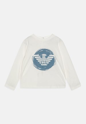 Camicia bianca a maniche lunghe con un design grafico blu che presenta un emblema di aquila circondato da accenti di pennellate circolari. Materiale in morbido cotone.