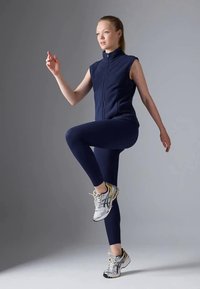 Tumši zils vējjakas stila veste ar rāvējslēdzēju pār saskaņotām legingiem, kopā ar sudraba un dzeltenām sporta apaviem. Modelis ir dinamiskā pozā uz pelēka fona.