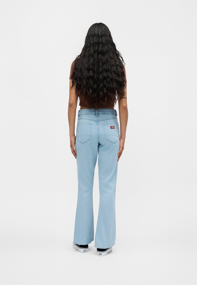 Frau mit langen welligen Haaren, die ein braunes kurzes Oberteil, hellblaue hoch taillierte Schlagjeans und weiße Turnschuhe trägt, steht mit dem Rücken zur Kamera.