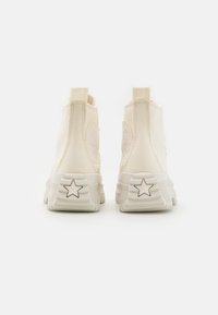 Höga sneakers i krämfärgad kanvas med texturerade ytor, med en rund emblem på sidan och stjärndetalj på hälen.