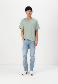 Lys grønn bomull t-skjorte med rund hals og trykt tekst på fronten, sammen med slitte lyseblå jeans og hvite joggesko.