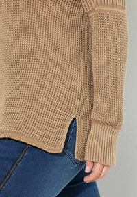 Beżowy sweter dziany z teksturowanym wykończeniem, ribbami przy mankietach i bocznymi rozcięciami. W połączeniu z ciemnoniebieskimi dżinsami.