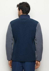 Giacca in pile con corpo blu navy e maniche grigie texturate, colletto alto e vestibilità rilassata. I dettagli delle cuciture aggiungono struttura.