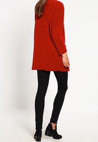 Punainen, teksturoitu ylisuuri takki pitkillä hihoilla, yhdistetty mustiin skinny-farkkuihin ja mustiin nilkkureihin, näkyy takaa.