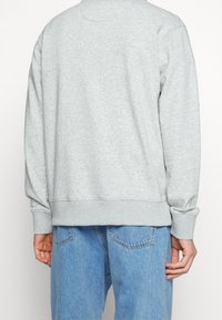 Persona vestida con un sudadera gris claro y jeans azul, mostrada de espaldas con las manos relajadas a los lados.