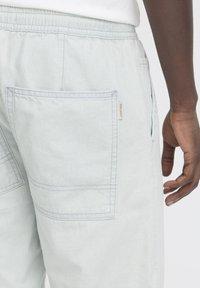 Pantaloni in cotone grigio chiaro con fascia elastica in vita, dotati di tasche posteriori e cuciture in contrasto. Vista ravvicinata del dettaglio della tasca e della mano.
