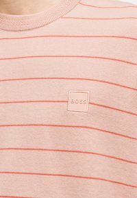 Ανοιχτό ροζ πλεκτό πουκάμισο με λεπτές οριζόντιες ριγέ σε σκουρότερο ροζ και τετράγωνο ταμπελάκι με την ανάγλυφη ένδειξη "BOSS" στο στήθος.