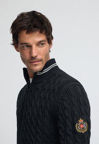 Svart stickad tröja med upphöjt flätmönster, hög krage, halv dragkedja och vit randig detalj vid halsen. Har en broderad logga på ärmen.