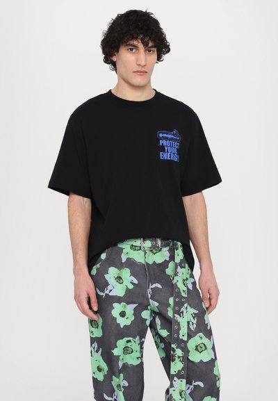Schwarzes kurzärmliges T-Shirt mit blauer grafischer Aufschrift "PROTECT YOUR ENERGY." Kombiniert mit grauen, floral gemusterten Hosen in Grün und Flieder.