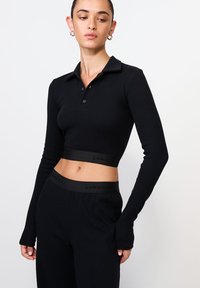 Fekete bordázott hosszúujjú crop top gombos nyakkal és tónusos rugalmas derékkal, amelyen a "LAMANIA" logó található.