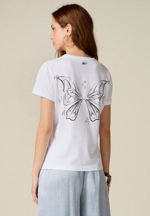 Jeune femme avec des boucles d'oreilles créoles portant un t-shirt blanc avec un motif d'ailes de papillon noires dans le dos, assorti à un pantalon bleu clair.