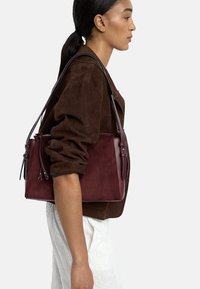 Bordowa torba z zamszu z skórzanymi akcentami, prostokątny kształt, regulowane paski i zamek błyskawiczny. Widoczna fakturowana powierzchnia.