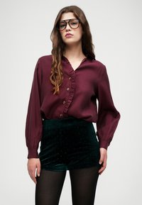 Burgundia volangidega pluus, millel on nuppude detailid, koos roheliste sametist lühikeste pükste ja täpilise mustriga, täiendab mustad sukkpüksid.