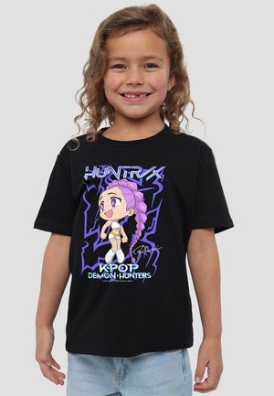 Smiling child wearing black T-shirt with purple anime character and "KPOP DEMON HUNTERS" text, standing against plain background.