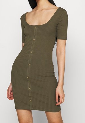 Femme portant une robe moulante côtelée de couleur vert olive avec des manches courtes et des boutons sur le devant, visible du cou jusqu'à mi-cuisse.