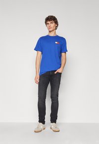 Mladý muž s kučeravými vlasmi oblečený v modrom tričku Tommy Hilfiger, čiernych skinny džínoch a béžových teniskách, stojaci s jednou rukou v vrecku.